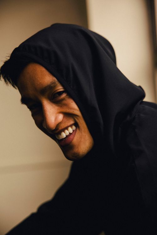 Close-up portrait of an adult man with dark hair wearing a black hooded sweatshirt, smiling with teeth showing against a light background.