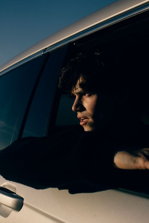 Close-up of a dark-haired young adult looking out of a white car window in shadowy lighting against a clear blue sky backdrop.
