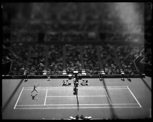 Monochrome shot of tennis player mid-swing on an outdoor court with a seated audience, capturing action and a vintage aesthetic.