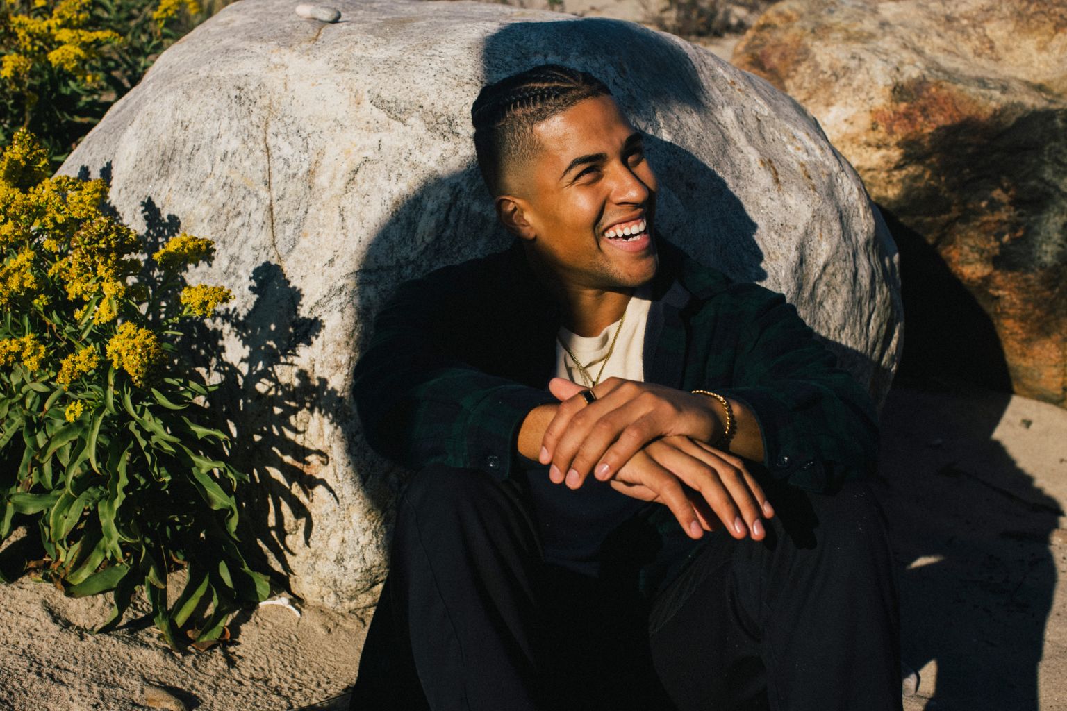 Smiling man poses next to a rock formation outdoors. The man sits and has his hands folded in his lap while glancing towards a sunlit space.
