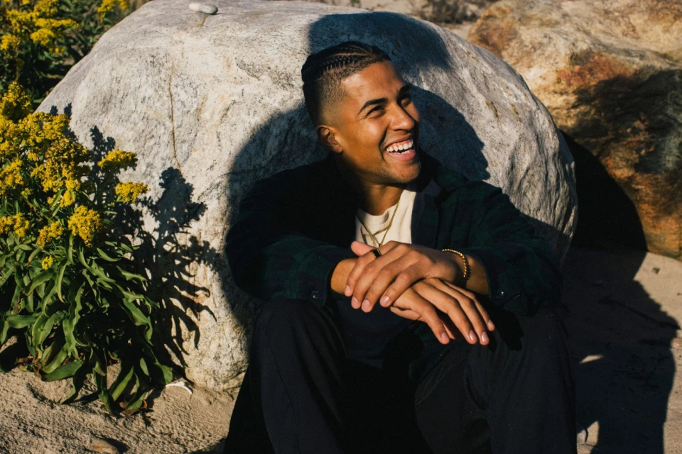 Smiling man poses next to a rock formation outdoors. The man sits and has his hands folded in his lap while glancing towards a sunlit space.