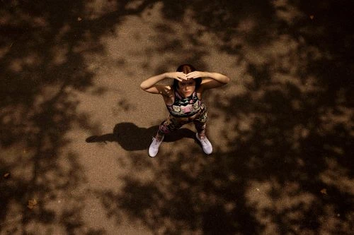 Overhead shot of woman in patterned athletic wear looking up with hands on head, on sun-dappled brown surface with tree shadows.
