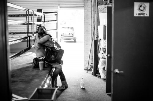 Black and white shot of two shirtless fighters embracing in a boxing ring. Focus is on figures, showcasing sportsmanship in stark tones.