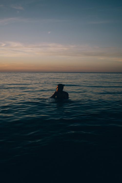 Person standing waist-deep in the calm ocean under a twilight sunset sky, captured by Nick Laham