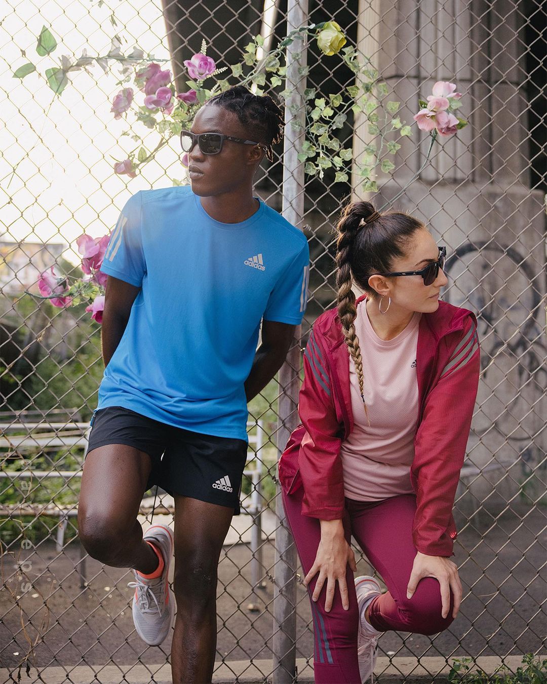 Two athletes, an adult male and female, in sport clothing and sunglasses, pose against a wire fence with climbing plants, looking away.