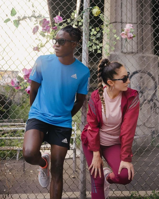 Two athletes, an adult male and female, in sport clothing and sunglasses, pose against a wire fence with climbing plants, looking away.