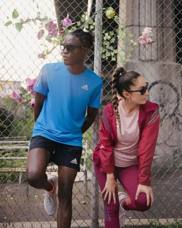 Two athletes, an adult male and female, in sport clothing and sunglasses, pose against a wire fence with climbing plants, looking away.