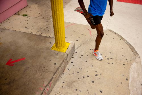 A man in athletic wear stretches his leg in an urban environment on a sunny day with yellow pillar and red arrow on the sidewalk.