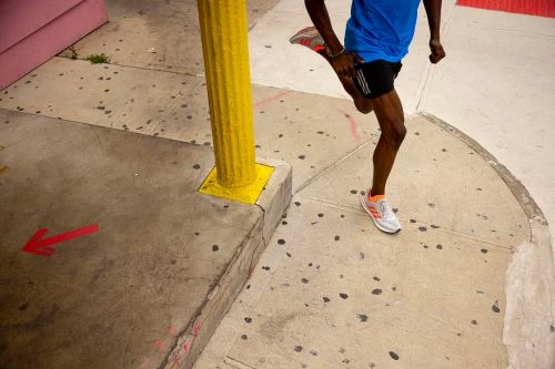 A man in athletic wear stretches his leg in an urban environment on a sunny day with yellow pillar and red arrow on the sidewalk.