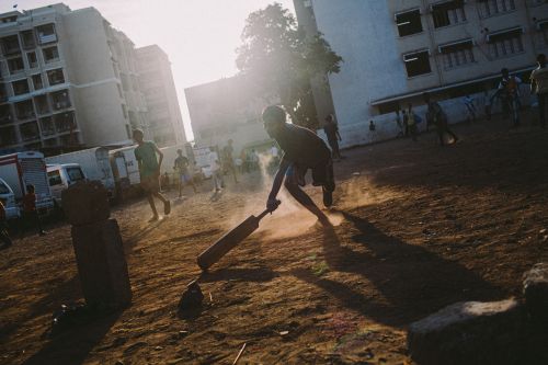 A youthful cricket player swings a bat on a dusty field, surrounded by other players and buildings in the background, capturing a sporty, urban moment.