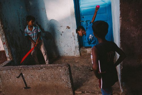 Three boys are playing cricket in front of a blue door on a sunny day, one holding a red bat, one holding a pink ball, one standing.