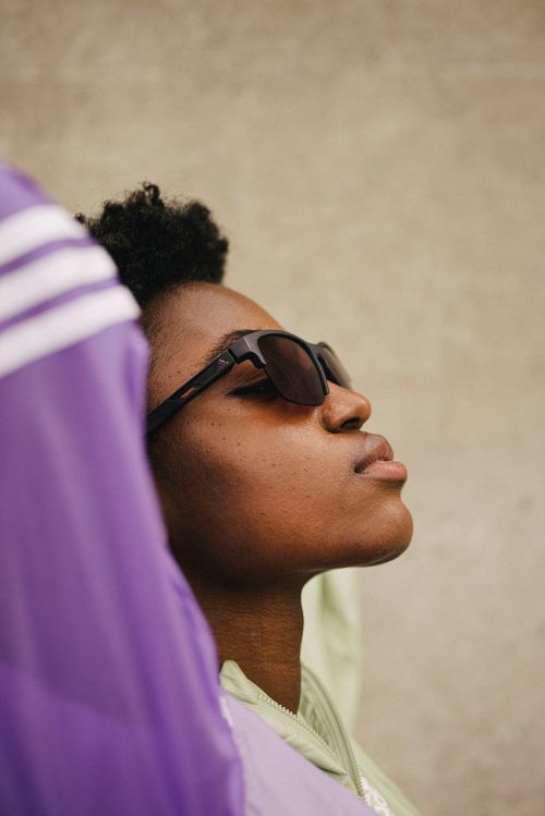 Close-up of a Black adult female in sunglasses, with an upward gaze, framed by blurred lilac and green apparel against a neutral background.