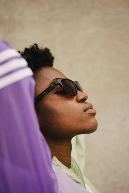Close-up of a Black adult female in sunglasses, with an upward gaze, framed by blurred lilac and green apparel against a neutral background.