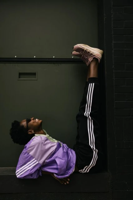 Woman in purple windbreaker and Adidas track pants poses upside down against a dark green wall, legs raised, smiling with sunglasses on.