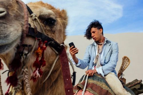 Man in a blazer riding a camel in the desert, holding up a smartphone, medium shot with a tan and blue color palette.