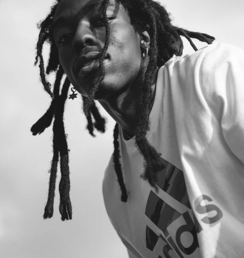 Monochrome portrait of an adult with dreadlocks wearing a branded t-shirt, showcasing a low angle shot and a bright, stark background.