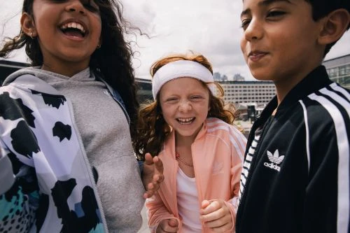Three smiling children outdoors, dressed in Adidas apparel, capturing a moment of joy and camaraderie against an urban backdrop.