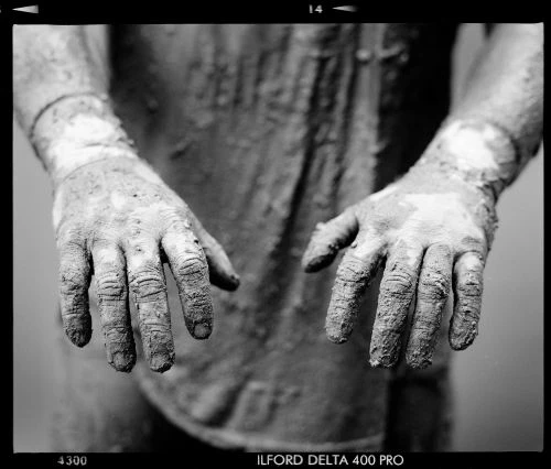 Black and white studio shot shows muddy hands extended, fingers spread, with dirty forearms and torso visible in the background.