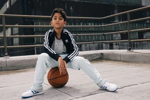 Young boy in Adidas jacket and sneakers poses with a basketball on a rooftop with metal railing and buildings in the background.