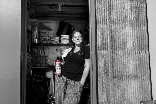 Monochromatic shot of a woman holding a beer can, standing in a doorway with corrugated metal siding, with cluttered shelves behind her.