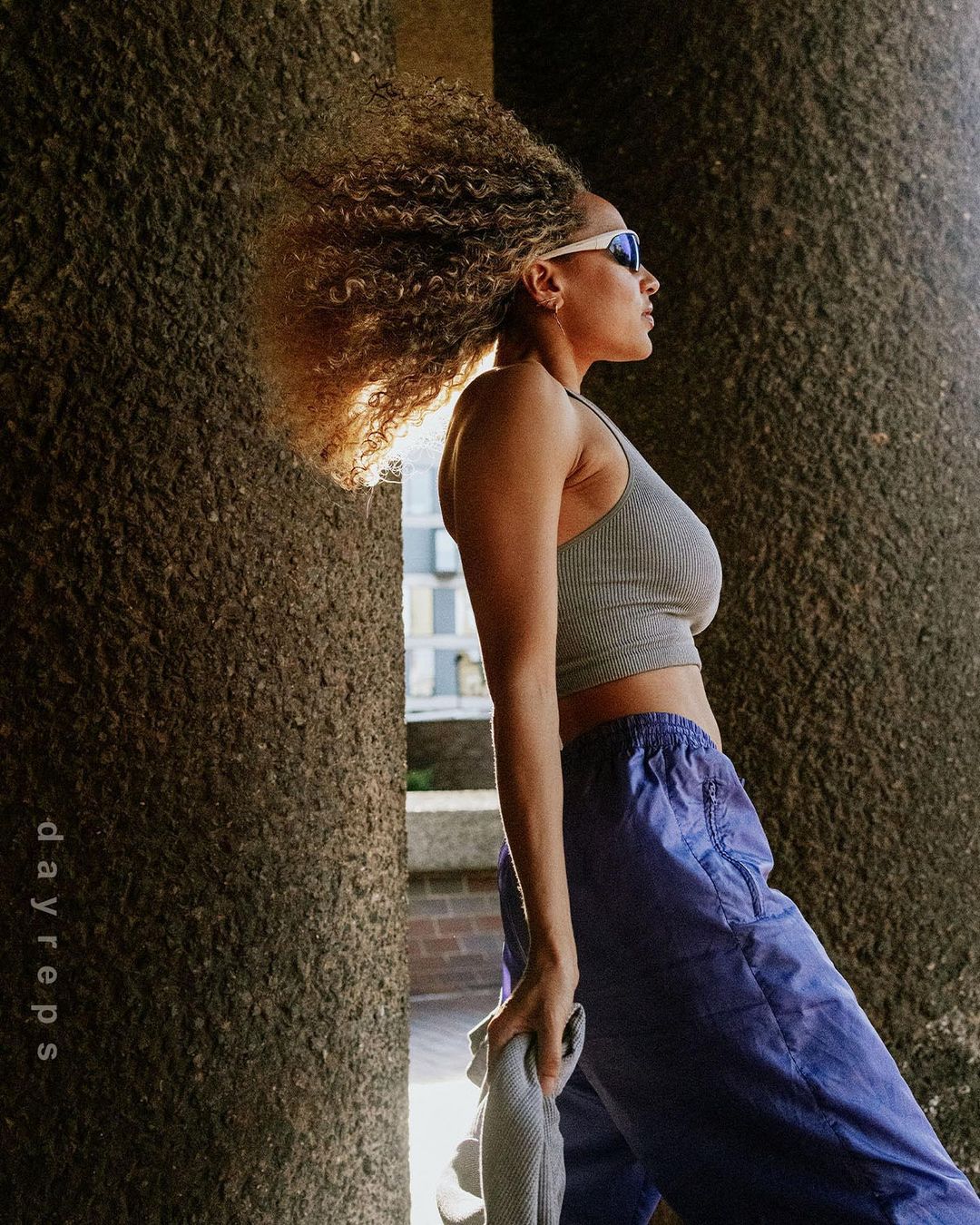 Woman with curly hair in athletic top and blue pants poses outdoors, with concrete architectural columns as a backdrop in the sunlight.