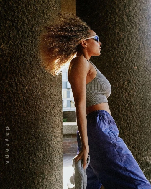 Woman with curly hair in athletic top and blue pants poses outdoors, with concrete architectural columns as a backdrop in the sunlight.