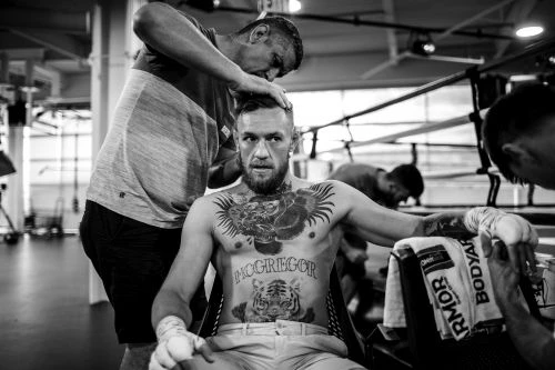 Black and white shot of a shirtless, tattooed Conor McGregor getting a haircut at a boxing ring, surrounded by people and training equipment.