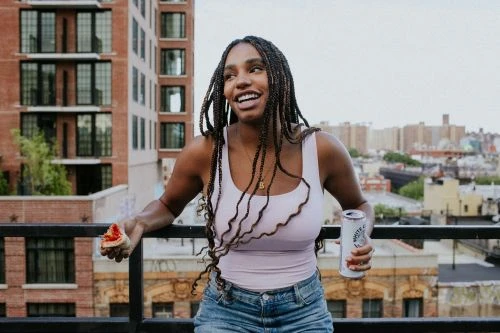 Smiling woman with braided hair, wearing a light purple top and jean shorts, stands on a rooftop holding a drink and pizza.