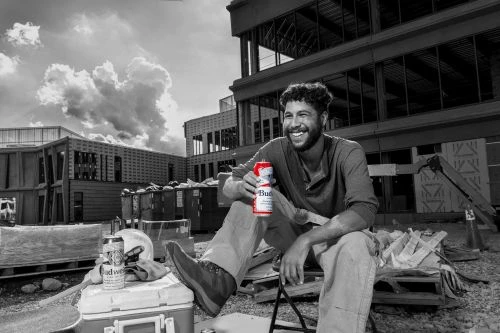 Man in work clothes smiles while sitting and holding a beer can on a construction site in black and white under a cloudy sky.
