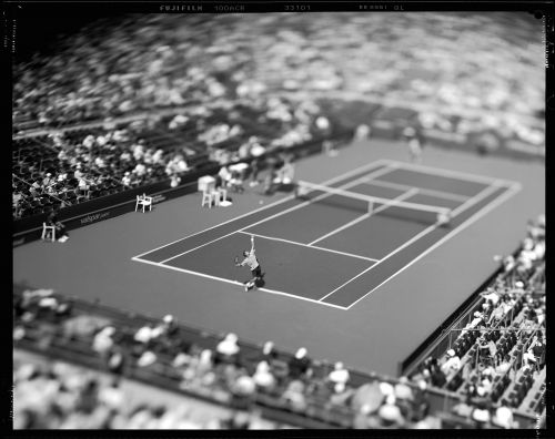 Monochrome action shot of a tennis player serving on a tennis court with spectators in the background. Sports and recreation photography.