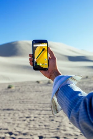 A hand holds a smartphone displaying a vibrant yellow floral pattern against a backdrop of sand dunes and a clear blue sky.