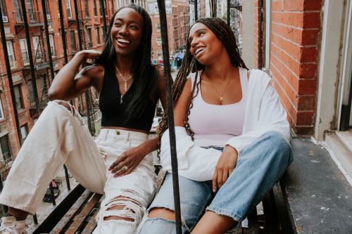 Two smiling young adult Black women sitting on a fire escape with brick buildings in the background, wearing casual tops and jeans.