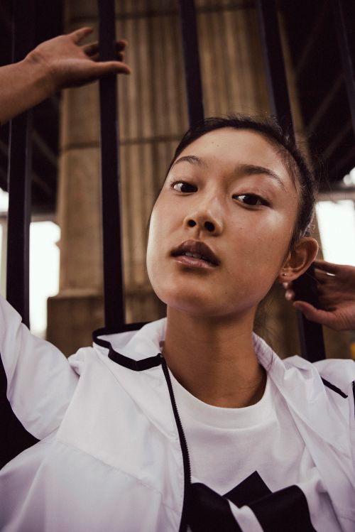 Portrait of an Asian woman in a white and black jacket, posing outdoors with hands up, in front of a dark metal structure.