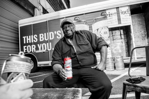 Black and white shot: African American man in hat and dark work clothes sits with a Budweiser can, smiling in an urban setting.