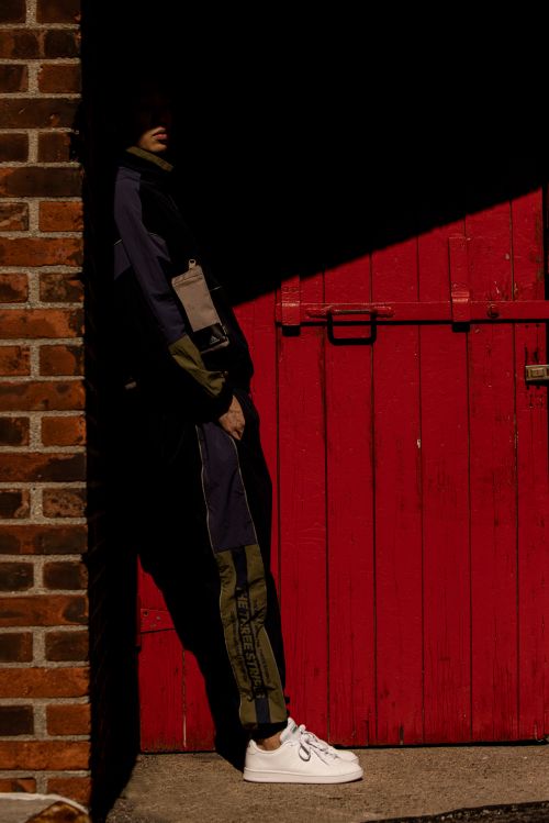 A person poses near a red door beside a brick wall, standing in shadows and wearing a track suit with white sneakers in an outdoor setting.