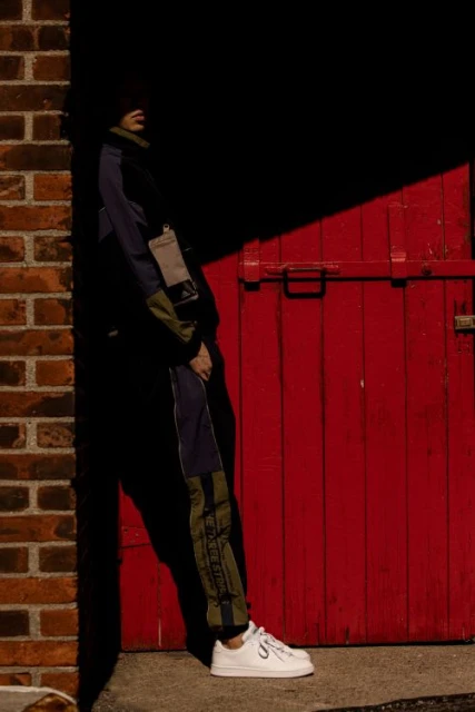 A person poses near a red door beside a brick wall, standing in shadows and wearing a track suit with white sneakers in an outdoor setting.