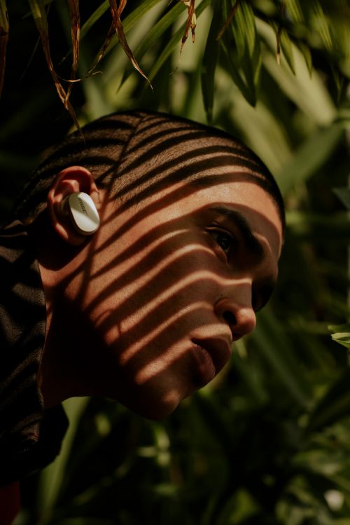 Close-up of a young adult wearing a wireless earbud, dappled in striped light and shadow, lush green foliage background.