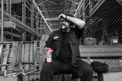 A man with a mustache sits outside on a pallet bench holding a beer, wearing a dark work shirt and dark pants, looking to the side.