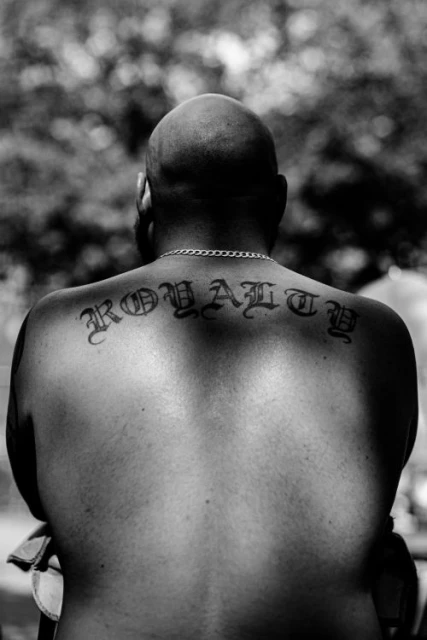 Monochrome portrait of a bald adult male with a tattoo on his back that says 'ROYALTY,' chain, and blurred foliage background.