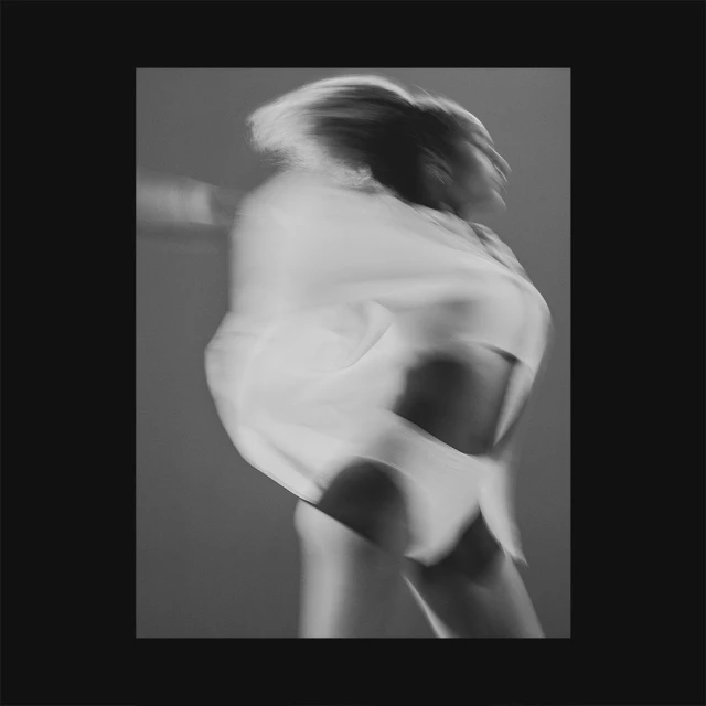 Blurred motion portrait of a person in a white garment against a neutral gray background, framed by a black border.