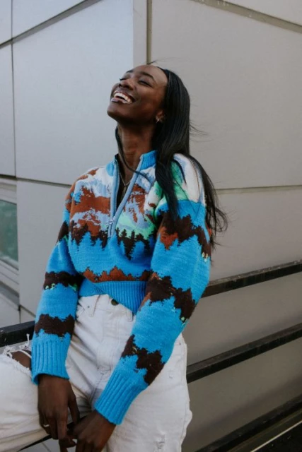 A smiling Black woman poses outdoors wearing a patterned sweater and ripped jeans, leaning against a railing with a building in the background.