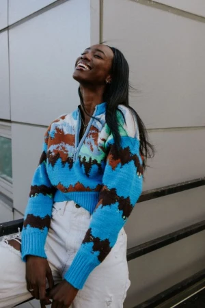 A smiling Black woman poses outdoors wearing a patterned sweater and ripped jeans, leaning against a railing with a building in the background.