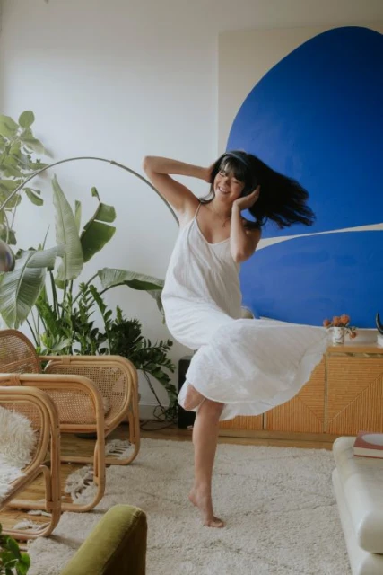 A cheerful adult woman dances barefoot in a sundress in a bright living room with plants, modern furniture, and an abstract painting.