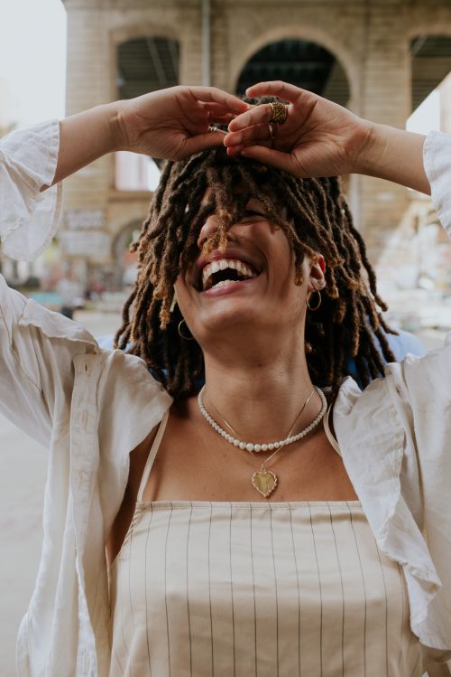 Laughing woman with dreadlocks raises arms in front of urban architecture, wearing jewelry and a striped top.