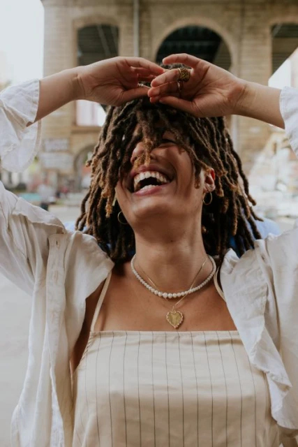 Laughing woman with dreadlocks raises arms in front of urban architecture, wearing jewelry and a striped top.