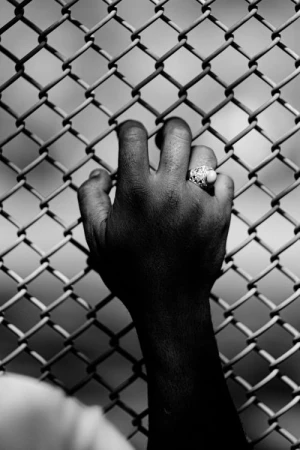 Monochrome image of an adult hand gripping a chain-link fence, showcasing a ring on the ring finger against a blurred background.