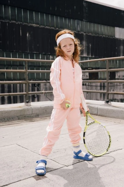 Young child in a peach-colored tracksuit and blue sandals standing on a rooftop with a tennis racket and tennis ball, looking at the camera.