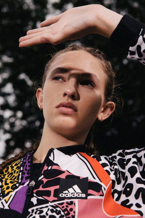Woman shields her face from sun wearing an Adidas color block jacket with braids in an outdoor setting with a blurred background.