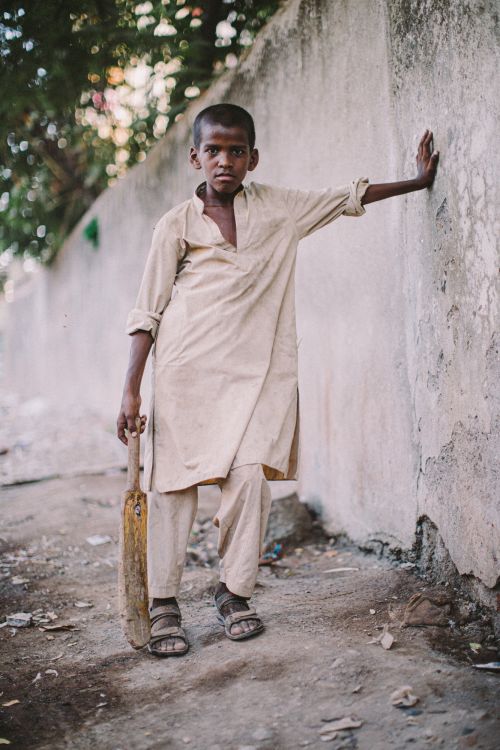 Young boy in beige shalwar kameez stands against concrete wall holding cricket bat, wearing sandals in an outdoor urban environment.