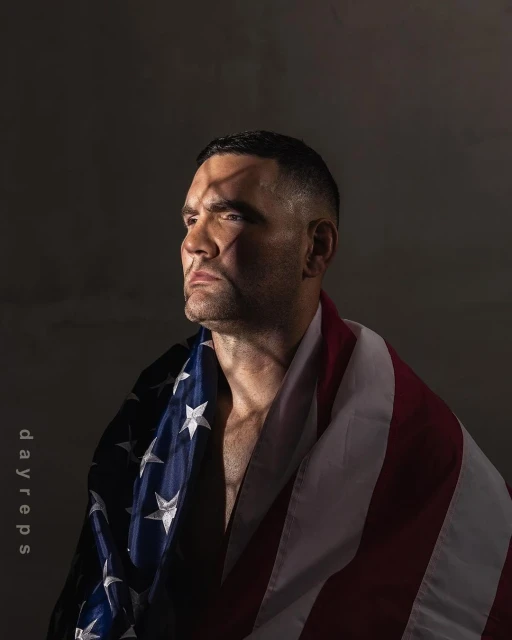 Adult male draped with an American flag looking off-camera in a low-key indoor portrait with hard rim lighting against a grey background.